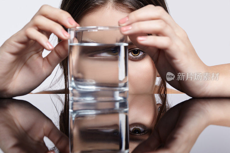 女孩把脸藏在盛有水的杯子后面。镜桌上年轻女子的光学扭曲肖像。