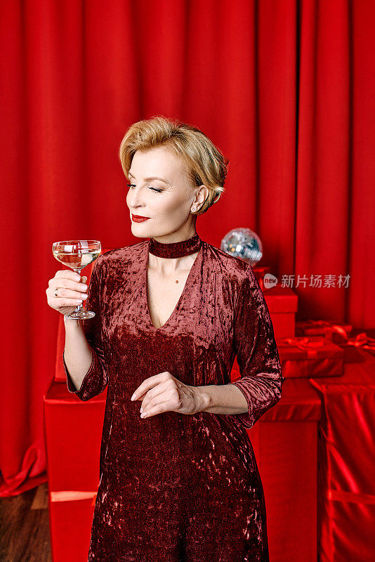 成熟时尚优雅的女人与一杯起泡酒与礼物在红色的背景。派对、时尚、庆典、反年龄概念