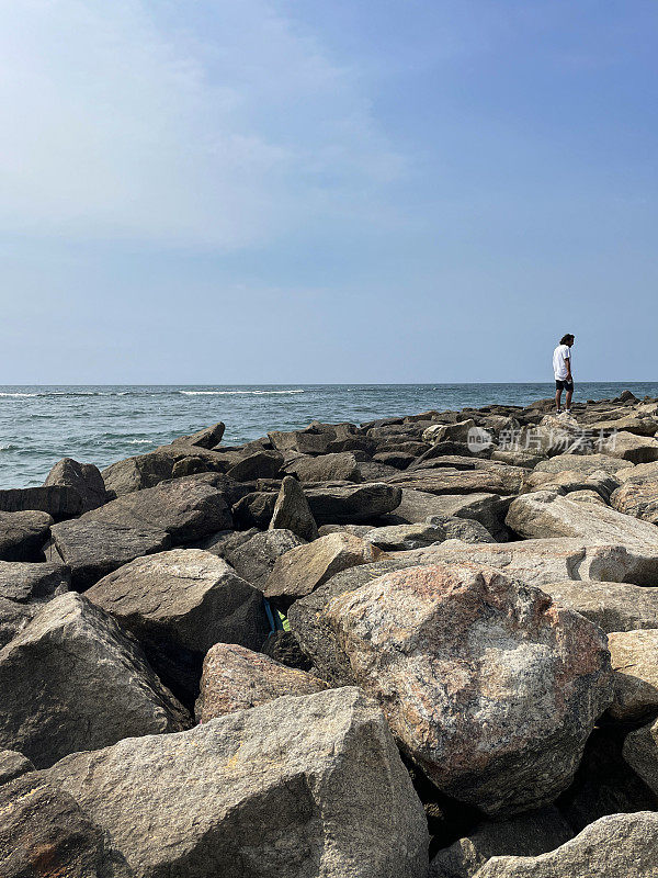 斯里兰卡，科伦坡，一名印度男子走过斯里兰卡海岸线上的人造岩石海防墙，海岸侵蚀抛石岩甲墙，海岸管理，大海，背景中的钓鱼竿