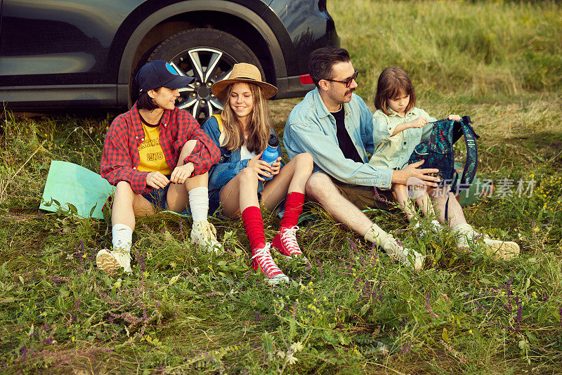 一家人，男人，女人和孩子坐在汽车旁边的健身matt上，在山上徒步旅行后休息。温暖的夏夜