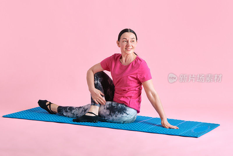 微笑，美丽，资深女性坐在健身matt，训练，伸展身体在粉红色的工作室背景