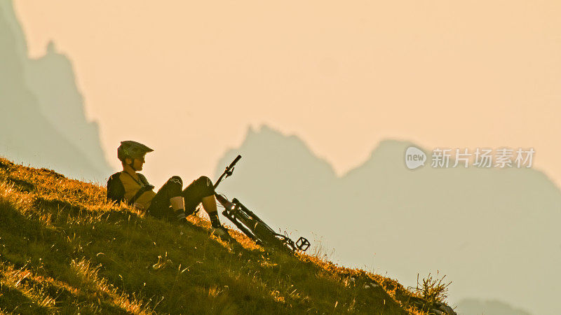 深思熟虑的男性山地自行车手在日落时在山上放松，面对晴朗的天空