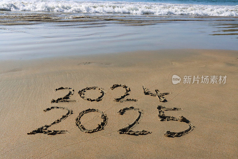 在阳光明媚的沙滩上用棍子在沙子上写2024年和2025年的特写图像，退潮时海水的边缘，在柔软的金色沙滩上写的数字，高架视图，南半球新年的概念