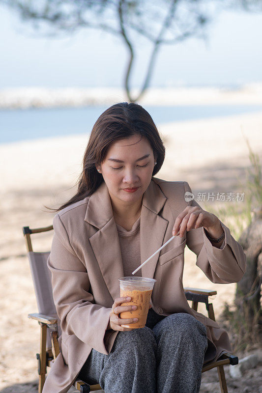 穿着棕色正装的亚洲妇女坐在海边的咖啡馆里，用吸管喝冷饮，以减轻牙痛。