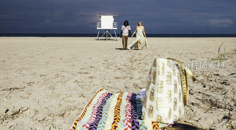 一对情侣留下毛巾和沙滩包，手牵着手走在田园诗般的海滩上