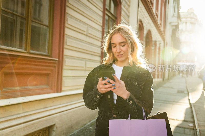 美丽的金发女孩走向相机，同时看着手机购物袋与太阳为背景-中镜头-日光