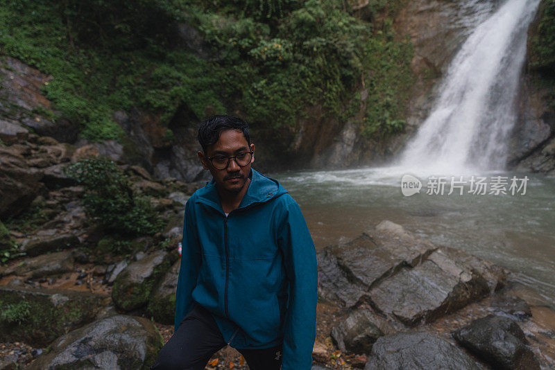 在热带雨林中，一名亚洲男子赤脚走在以瀑布为背景的石头上