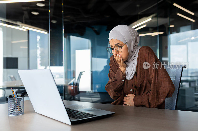 戴着头巾在办公室里工作的生病的女商人，穆斯林妇女恶心和胃痛，呕吐的妇女，坐在桌子上用笔记本电脑呕吐