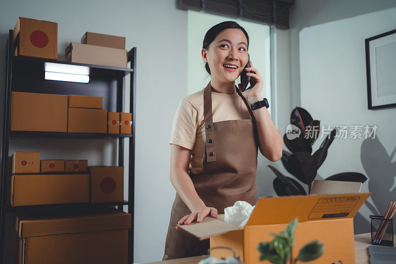 亚洲女性在线卖家用智能手机打电话给客户确认订单，并包装产品准备发货。