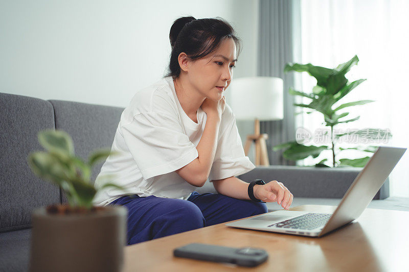 一名亚洲女性坐在家里的沙发上使用笔记本电脑，身体疼痛。