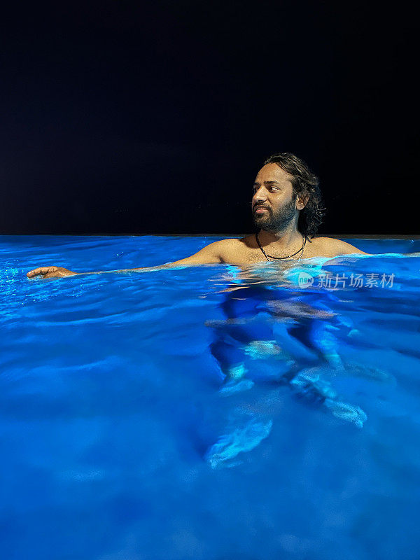 画面中，印度男子肩膀深埋在无边泳池的水里，靠着玻璃墙泳池边，蓝色的水下灯光，夜空背景，聚焦前景