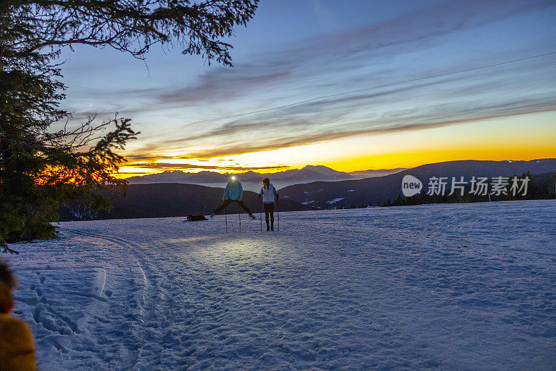 黄昏时分，两名女滑雪者手持滑雪杖在雪坡上滑雪