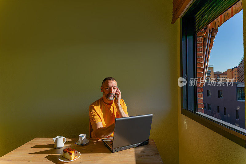 一位中年男子一边打电话，一边看着放在木桌上的笔记本电脑的屏幕，桌上还有一杯咖啡和一块蛋糕。