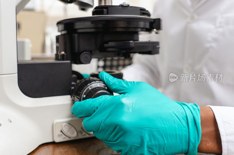 在生物学研究中心，年轻的成年科学家戴着防护手套用旋钮调节光学显微镜的图像聚焦，专业人员使用实验室设备进行研究和开发