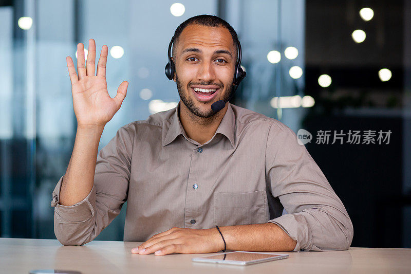 微笑快乐的非洲裔美国人戴着耳机进行视频通话，在现代办公室咨询客户，在网上商店支持工作，在相机前挥手，镜头视图