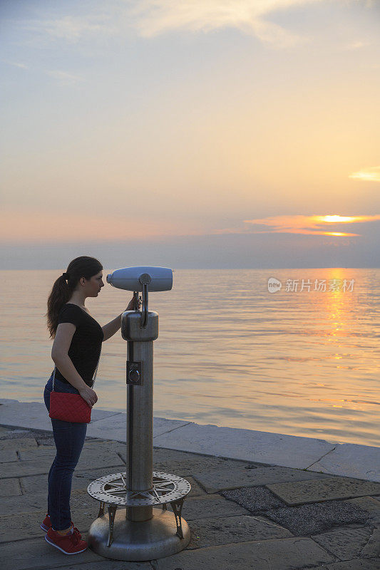 年轻女子透过投币双筒望远镜眺望海洋漫步海上日落