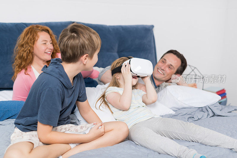 女儿戴着虚拟现实数字眼镜与父母和兄弟坐在床上，家人玩得很开心