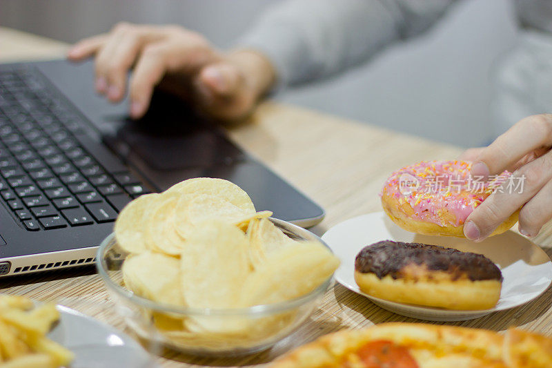 一个人在电脑前工作，吃快餐。不健康食品:汉堡，酱汁，土豆，甜甜圈，薯条。