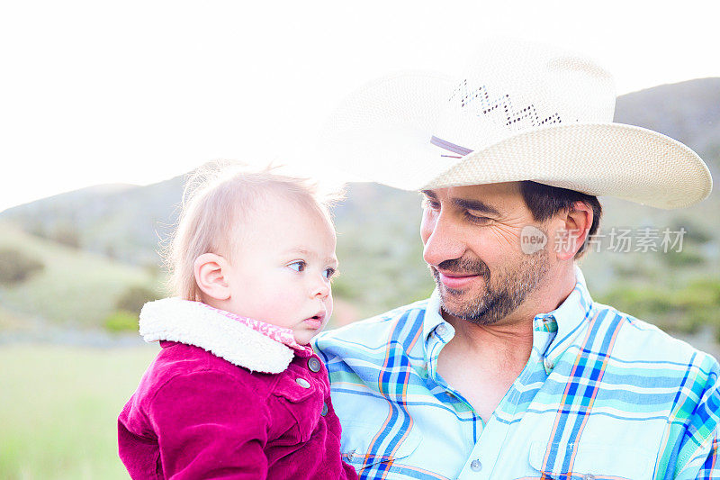 西方生活方式的父亲在牛仔帽慈爱地抱着他的蹒跚学步的女儿