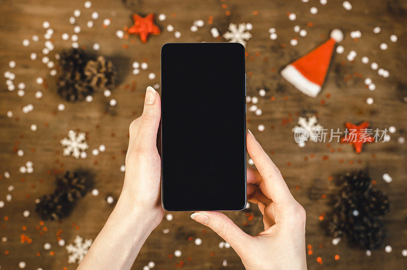 女性手握智能手机与oled显示屏上的木制背景与圣诞礼物雪花和雪花。新年快乐，帽子上的圣诞老人手机，平铺构图俯视图。