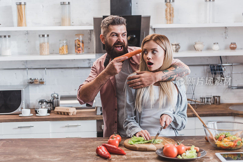一个滑稽的男人把胡萝卜放在女人的头旁边，而受惊的女孩正在做晚餐
