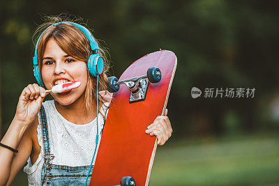 美丽的年轻女子与滑板在一个可爱的晴天在公园，听音乐和吃新鲜的冰淇淋