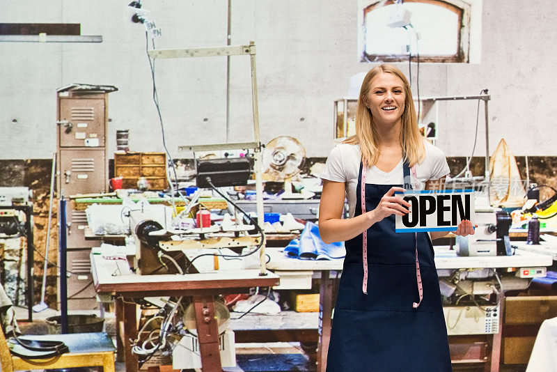 年轻的女企业家穿着围裙，拿着开着的牌子，用缝纫机在布料店里缝纫