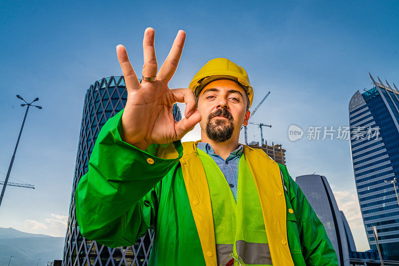 年轻的工程师戴着黄色安全帽和绿色防护服在现代化的商业建筑和建筑工地前看着摄像机，并显示OK手势。镜头光晕。低角度的观点