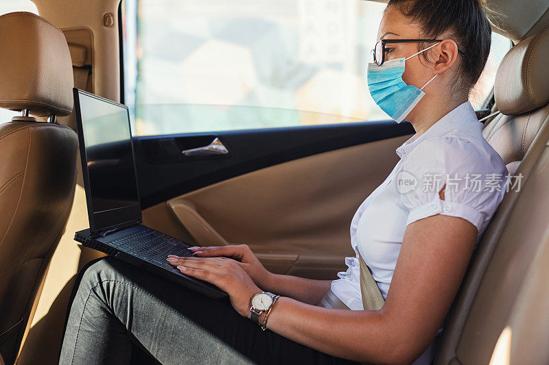 年轻美丽的女商人戴着保护面罩，坐在车里用笔记本电脑工作