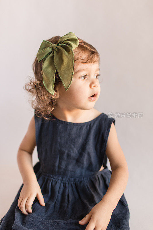 两岁蹒跚学步的小女孩穿着海军蓝亚麻太阳裙和橄榄绿头发蝴蝶结，看着窗外