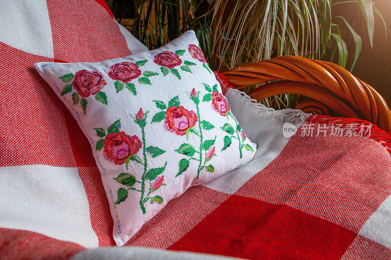 白色枕头上的玫瑰刺绣，躺在扶手椅上，铺着红白相间的毯子，背景是植物。舒适房间的内部