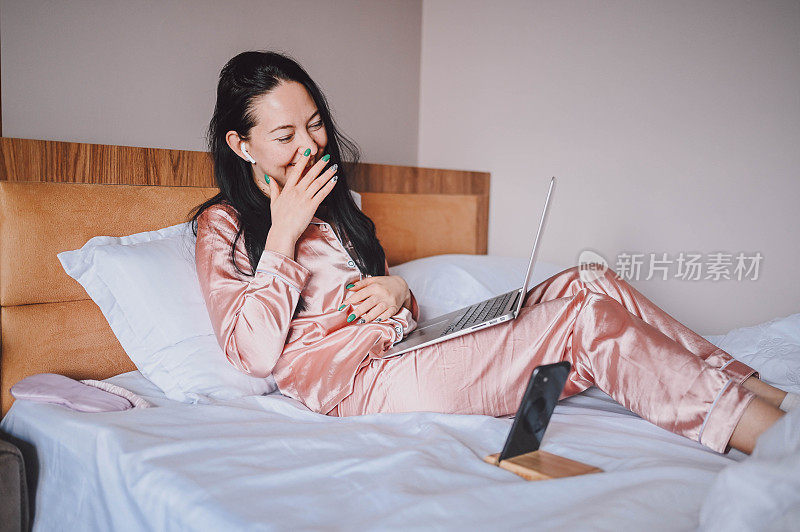 年轻快乐的黑发女子，戴着无线耳机，穿着粉色丝绸睡衣躺在床上，用智能手机和笔记本电脑，进行在线视频聊天。Blogger直播早间新闻