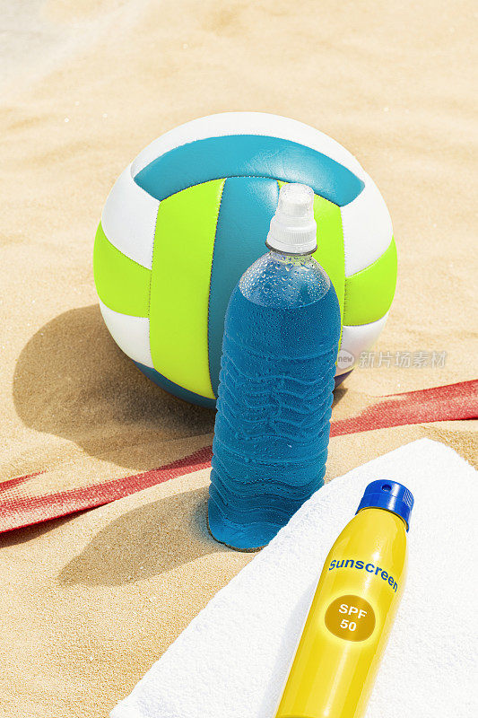 沙滩排球坐在旁边，毛巾上放着一瓶防晒霜，沙滩上放着运动饮料