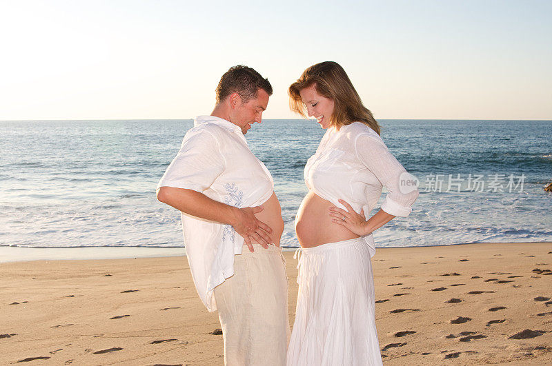 美丽的怀孕夫妇在海滩上比较肚子