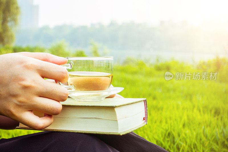 女人坐在草地上，手里拿着茶和书