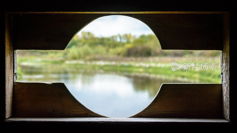 从一个观鸟的木制掩体通过一个开口与圆形切口适合长焦镜头从一个池塘。清晰的前景(窗口)，模糊的背景(风景)