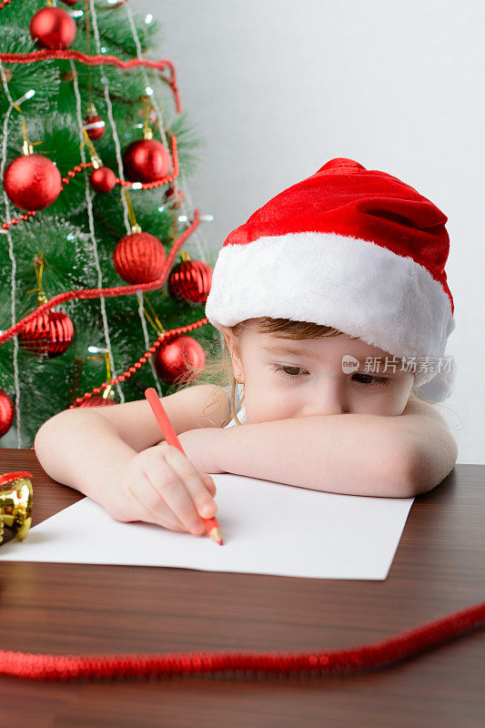 给圣诞老人写信的概念。可爱的小女孩拿着一支笔坐在圣诞树旁的桌子旁
