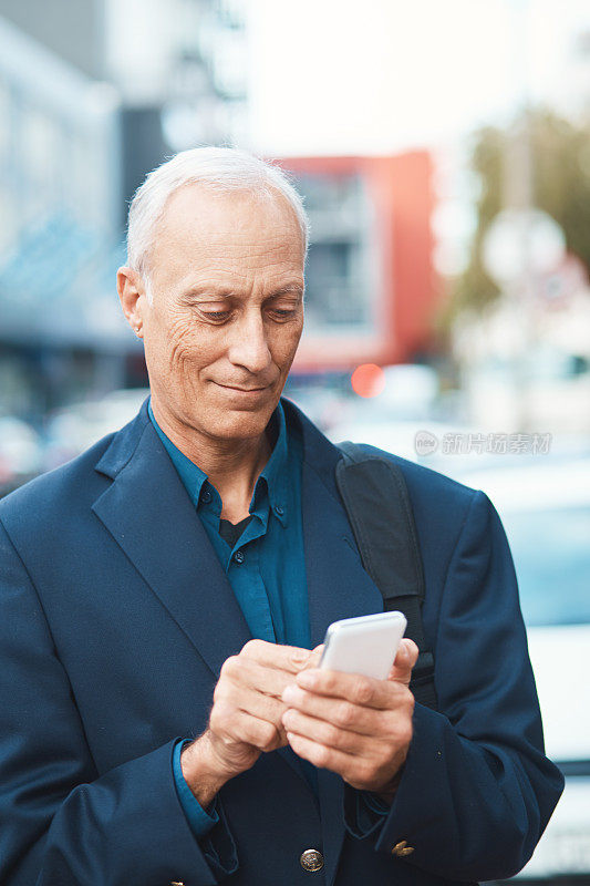 成熟的商人微笑着，在城市街道上用智能手机发短信