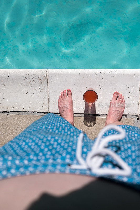 视点拍摄，俯视着一个穿着蓝色泳裤站在室外游泳池边上的脚边喝酒的男人。一个穿着泳衣的白人站在泳池边。