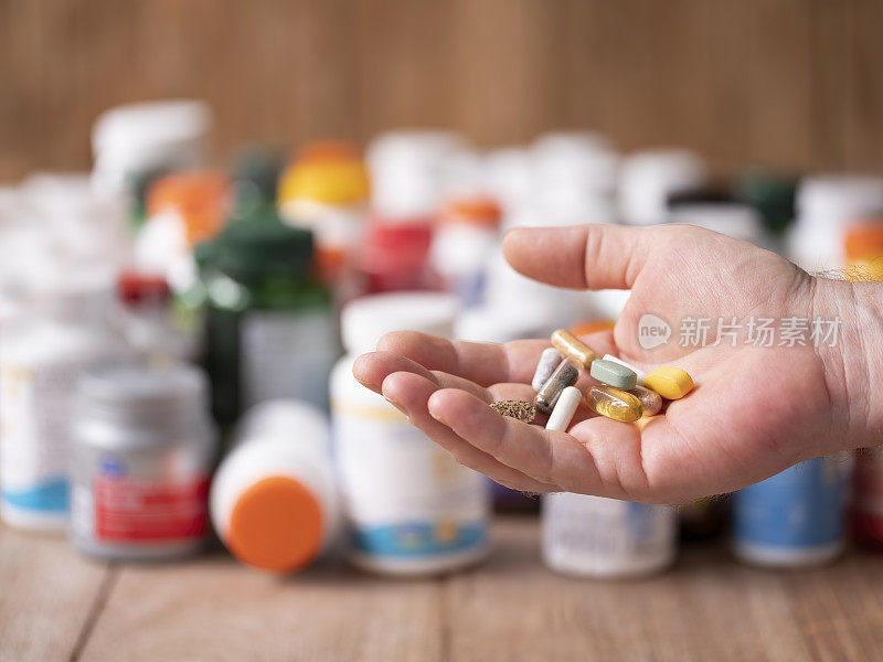 许多营养保健品和维生素胶囊和药片在食用前拿在手里，许多五颜六色的食品保健品和维生素瓶子在一张木桌的背景中。