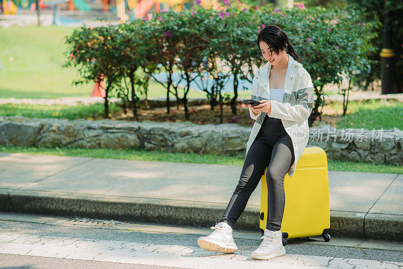 亚洲华裔少女玩她的智能手机，坐在滚动的行李，等待出租车司机来接她