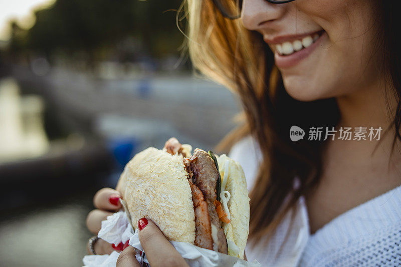 一个年轻的棕色头发的女人一边吃着三明治一边走在街上——一个年轻的时髦女孩走在街上，享受着她的三明治