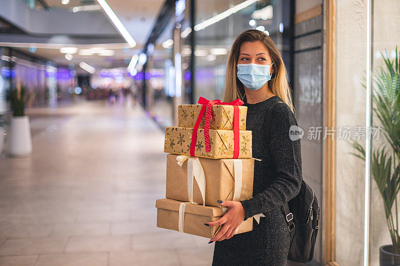 新冠肺炎疫情期间，年轻美丽的女孩在商场享受圣诞购物。她戴着防护口罩，以防止冠状病毒感染。