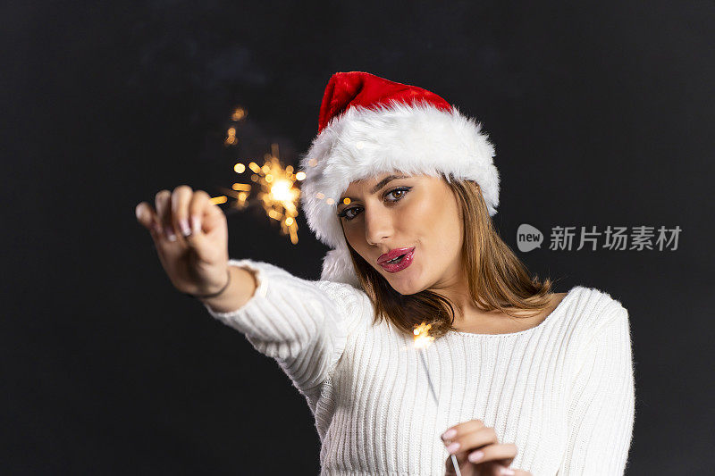 美丽的年轻女孩与圣诞老人帽拿着燃烧的烟花在圣诞节在黑色的背景
