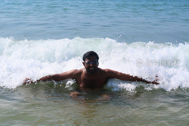 一名印度男子在印度果阿的帕洛伦海滩度假，坐在印度洋上溅水花，戴着护目镜，伸开双臂溅水，看起来很开心
