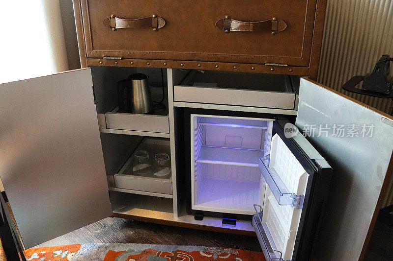 开放的酒店橱柜展示小集成冰箱旁边的置物柜与电热水壶和饮水杯，打开冰箱门展示空货架和室内光，重点在前景