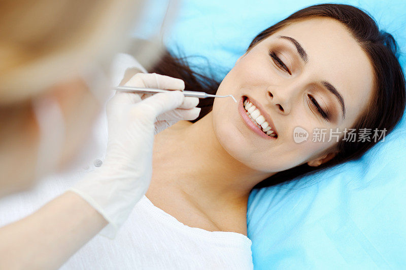 年轻女性病人前往牙科诊所。美丽的女人与健康的直白牙齿坐在牙科椅张开嘴在口腔检查期间，而医生在牙齿工作
