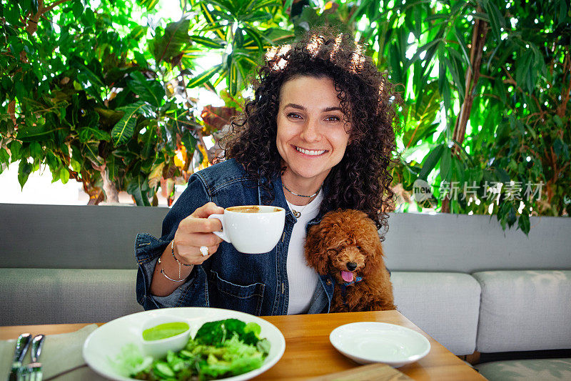 美丽卷曲的黑发女人的肖像，带着宠物在素食餐厅享受植物性蔬菜餐，环保的室内装饰着郁郁葱葱的绿色植物