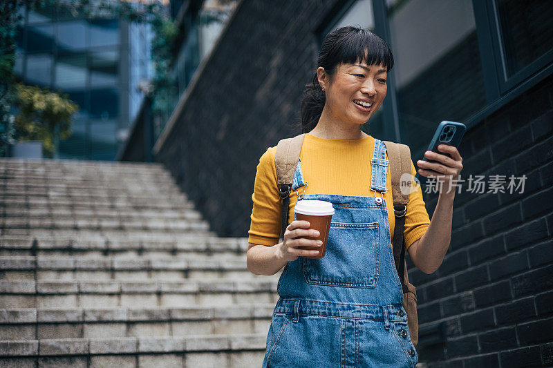 一名日本女子拿着智能手机和一杯咖啡，看着手机屏幕，背景是一栋办公楼