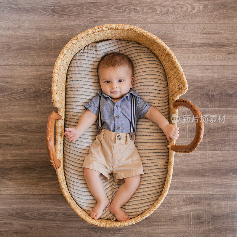 一个22周大的男婴穿着蓝色背带裤，躺在一个舒适的奶油色条纹棉毯在海草摩西篮子里